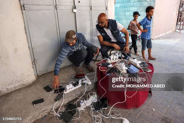 People sit by phone chargers connected to a communal group of electric socket hubs outside a closed pharmacy in Rafah in the southern Gaza Strip on...