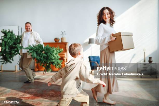 middle-aged mom and dad and child son bring box of christmas decorations and christmas tree into bright living room, preparing to decorate apartment for christmas - milestone stock pictures, royalty-free photos & images