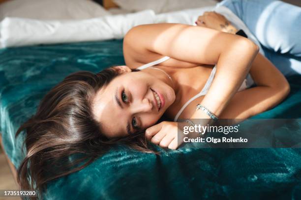 morning laziness in cozy bedroom. smiling brunette woman resting in her bed on weekends. woman lying on side and look at camera - brunette woman bedroom stock-fotos und bilder