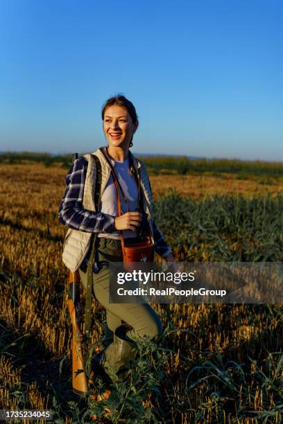 young female hunter cheerfully standing in a wheat filed, while holding binoculars and carrying a shotgun - hunting rifle stock pictures, royalty-free photos & images