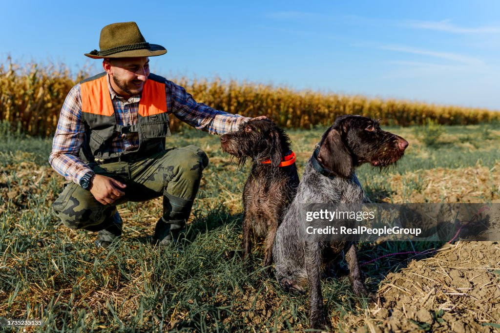 Young male hunter crouching next to his two trusty hunting dogs and petting them