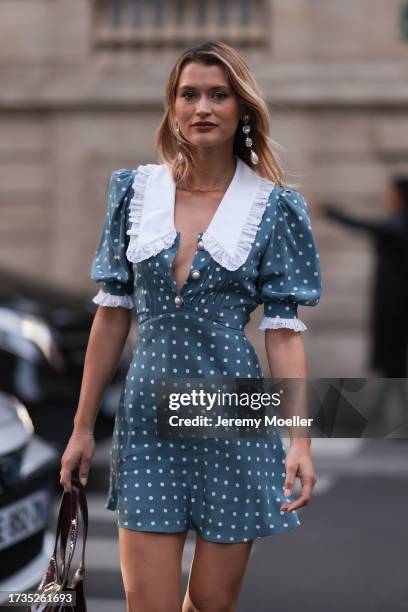 Chloe Lecareux is seen wearing a blue satin dress with padded shoulder sleeves, white collar, black patent handbag, and long pearl earrings during...