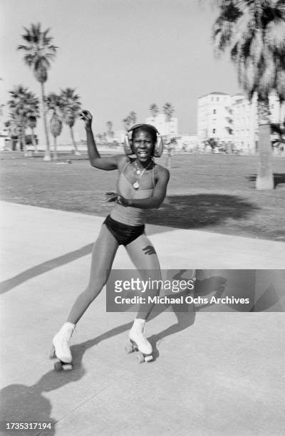 Young woman wearing a vest with spaghetti straps, shorts, and a pair of headphones, roller skating on the beachfront of Venice Beach, in the Venice...