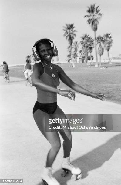 Young woman wearing a vest with spaghetti straps, shorts, and a pair of headphones, roller skating on the beachfront of Venice Beach, in the Venice...