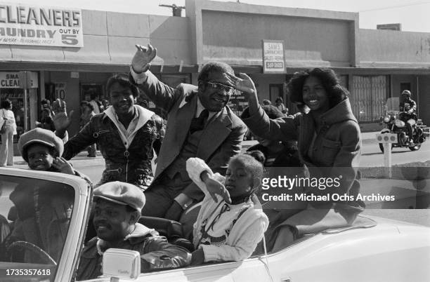 American politician Bill Greene, California State senator, waving as he sits with children in an open-top Cadillac during the Watts Christmas Parade,...