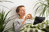 Throughout the day, a middle-aged woman with short hair lounges in bed, glued to her tablet screen and munching on a sandwich. Indolence, passivity. Bed rotting