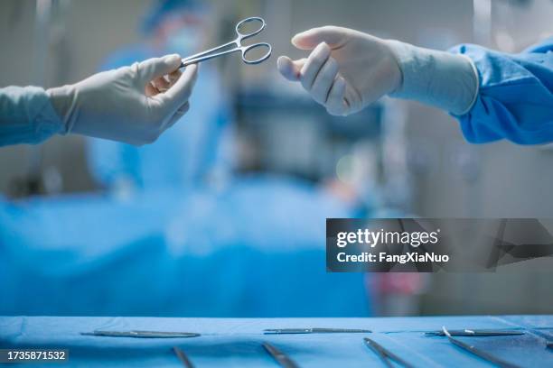 hands of nurse passing medical tool to doctor wearing protective workwear in medical surgery operation room in hospital with patient incidental people background - surgical glove stock pictures, royalty-free photos & images