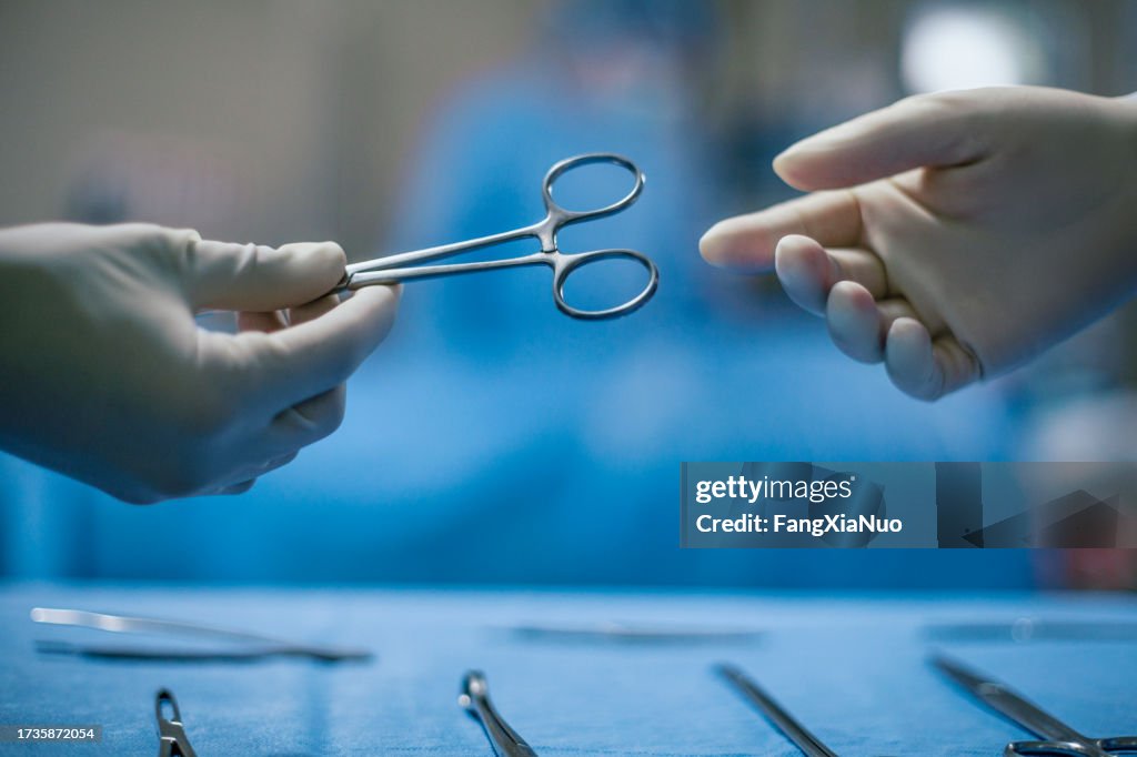 Mani dell'infermiera che passa lo strumento medico al medico che indossa indumenti da lavoro protettivi nella sala operatoria della chirurgia medica in ospedale con lo sfondo accidentale delle persone del paziente