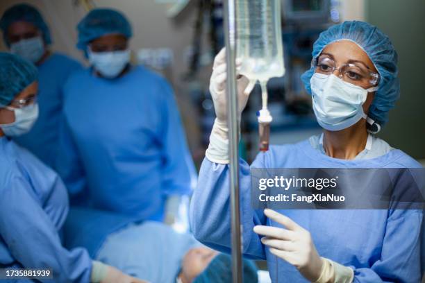 multiracial hispanic black ethnicity woman nurse doctor checking iv drip medication for patient in surgery operating room in hospital wearing protective workwear with colleagues anesthesiologist - anesthesioloog stockfoto's en -beelden