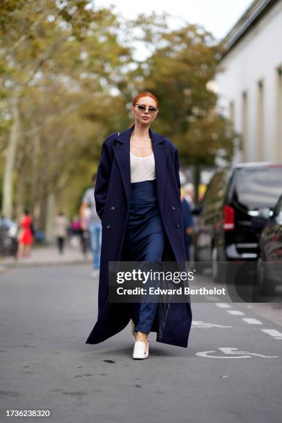 Guest wears sunglasses, a navy dark blue long coat, a white low neck tank top, blue pants, white shoes, outside Anrealage, during the Womenswear...