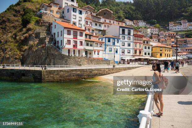 chinese young woman wearing sunglasses and carrying a rucksack stands next to a railing looking away with the colourful town of cudillero by the sea on the background in asturias, northern spain - asturias stock pictures, royalty-free photos & images