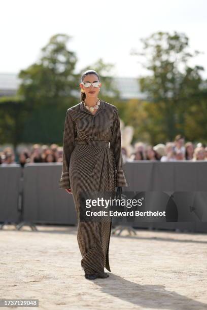 Bettina Looney wears silver mirror sunglasses, a necklace made of items designed as sea shells, a brown and black gathered long dress with checkered...