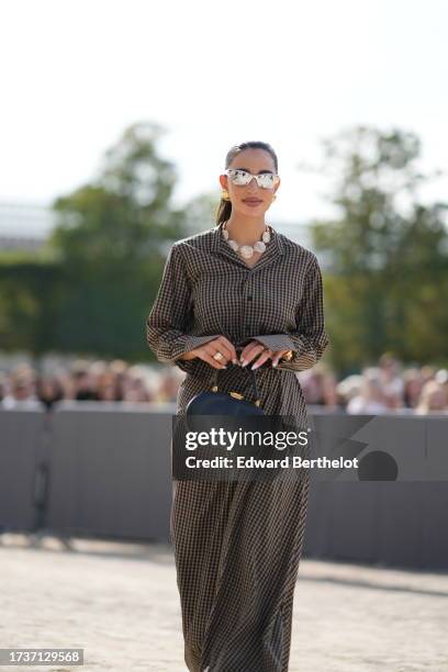 Bettina Looney wears silver mirror sunglasses, a necklace made of items designed as sea shells, a brown and black gathered long dress with checkered...