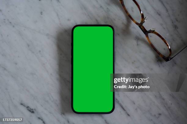 a flat lay of a mobile phone with blank green screen on a marble table background with an eye glasses. - green screen stock pictures, royalty-free photos & images