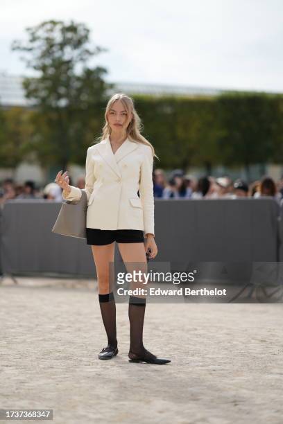 Anoushka Gauthier wears sunglasses, a white double breasted oversized blazer jacket, black shorts, knee high tights, black shiny shoes, a gray bag,...