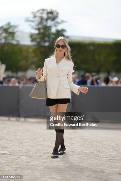 Anoushka Gauthier wears sunglasses, a white double breasted oversized blazer jacket, black shorts, knee high tights, black shiny shoes, a gray bag,...