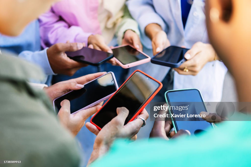 Young group of people standing in circle using mobile phones outside. Unrecognizable teen friends watching social media content on smartphone app. Technology lifestyle concept.