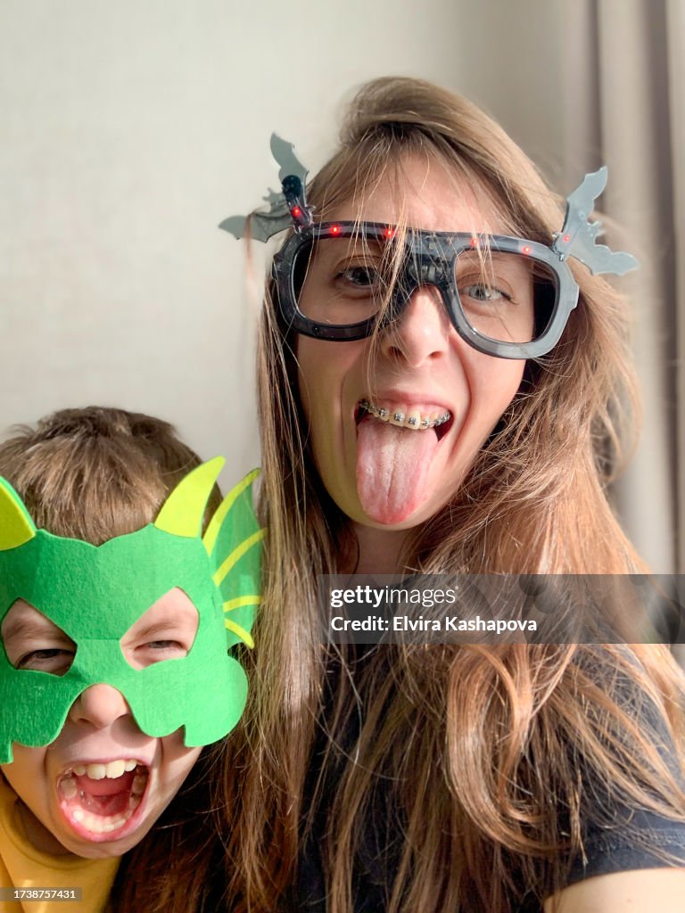 Mother and son show tongue in masquerade halloween masks and take selfie. Mom in black glasses, son in a dragon mask making faces