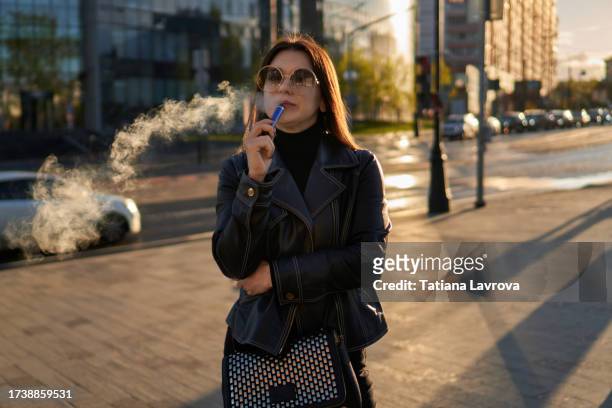 attractive young female smoking in the street. lady of a millennial generation vaping a vaporizer. fashion photography background with space for design - vape stock-fotos und bilder