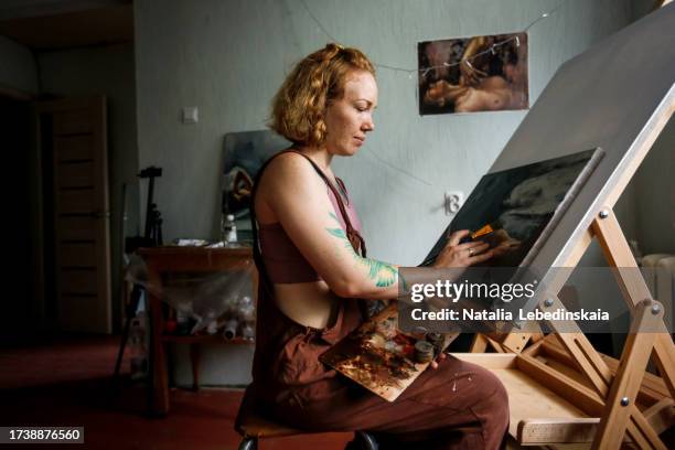 canvas creativity: youthful red-haired woman engages in painting on easel with paints in her workshop. dark style - malerleinwand stock-fotos und bilder
