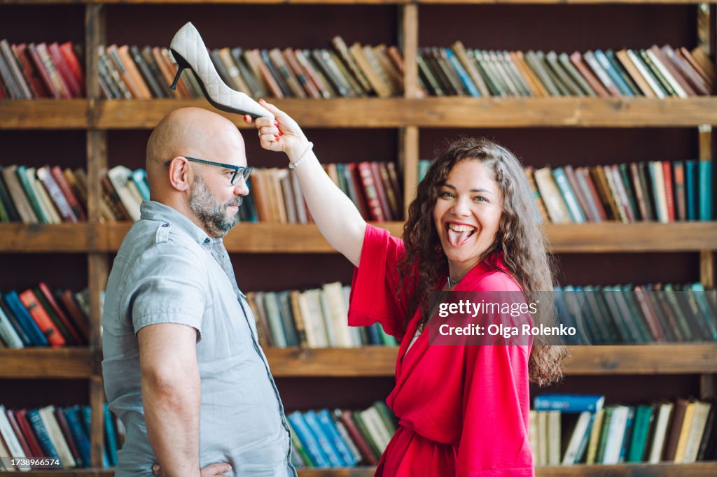 Henpecked husband. Laughing brunette woman holding shoe heels above man head in library. Obedient husband issues