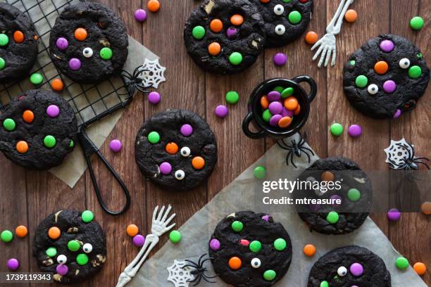 image of homemade halloween, white chocolate chip, black cocoa cookies on cooling rack, decorated with pink, green and orange candy coated chocolate sweets, plastic skeleton arms, webs, plastic spiders, cauldron, wood grain background, elevated view - cauldron stock pictures, royalty-free photos & images