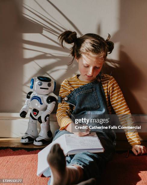 a child sits on the floor, writing in a notepad. an educational robot toy sits beside her. - brain thinking goal setting stock pictures, royalty-free photos & images