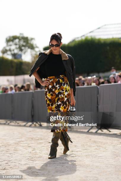 Guest wears sunglasses, an aviator jacket, a black top, a yellow and brown floral print midi skirt, brown leather pointed boots, outside Dior, during...