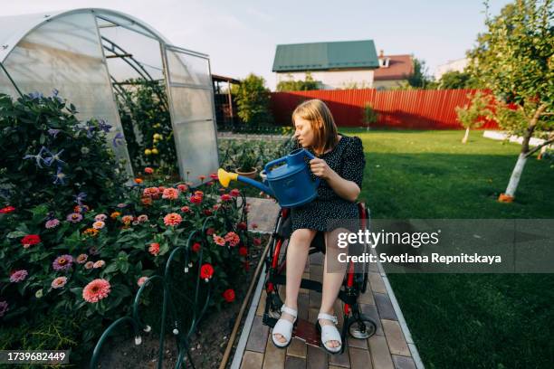 girl with cerebral palsy in a wheelchair waters flowers from a watering can in her garden - world kindness day stock pictures, royalty-free photos & images