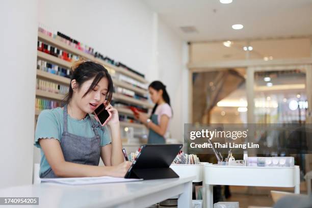una joven asiática está haciendo varias cosas a la vez en su salón de uñas. recibe una llamada de un cliente para una cita y, al mismo tiempo, utiliza una tableta digital para actualizar su calendario de trabajo. - salón de belleza para manicura fotografías e imágenes de stock