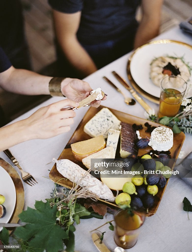 A table laid with a white cloth and place settings seen from above. An organic cheese board with soft and hard cheeses and figs. Two people sitting at the table.