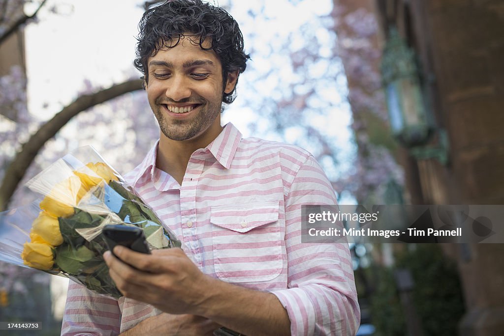 City life. A young man in the park in spring, using a mobile phone.  Holding a bunch of yellow roses. 