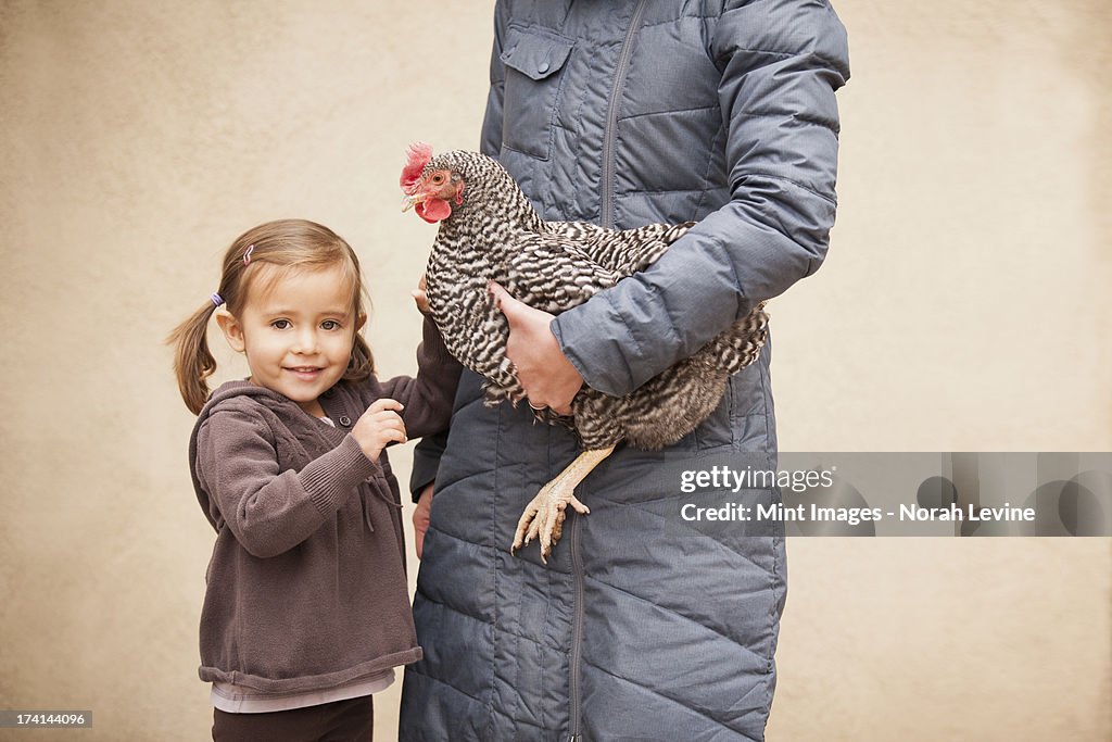 A woman in a grey coat holding a black and white chicken with a red coxcomb under one arm. A young girl beside her holding her other hand