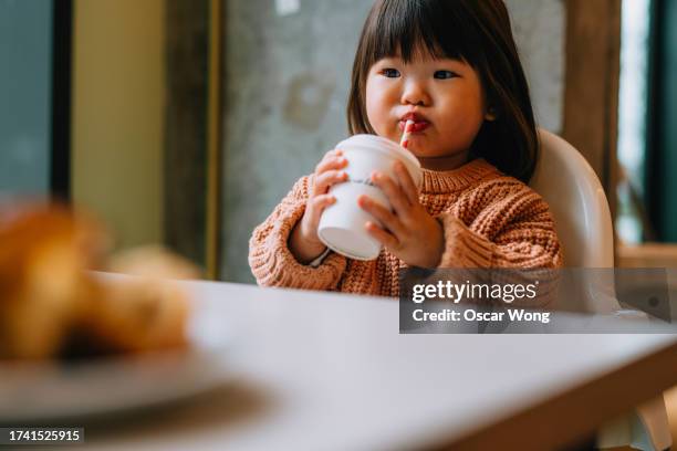 cute asian toddler girl holding a cup and drinking babyccino from a paper straw at cafe - toddler milk stock-fotos und bilder