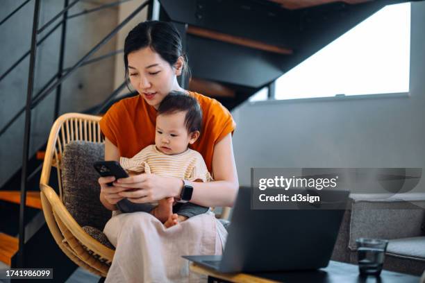 young asian mother working at home with smartphone and laptop while taking care of her baby girl. working mother. parents working from home with kids. managing home finances. multi-tasking working mother managing work life and family life at home - asian girl doing online shopping ストックフォトと画像