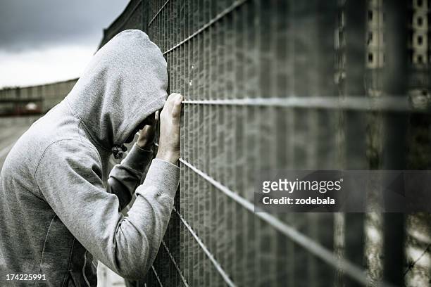 lonely mann mit kapuze auf einen zaun lehnend - prison stock-fotos und bilder