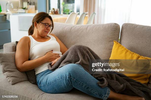 mature woman suffering from stomach cramps on the sofa at home - irritable bowel syndrome stock pictures, royalty-free photos & images