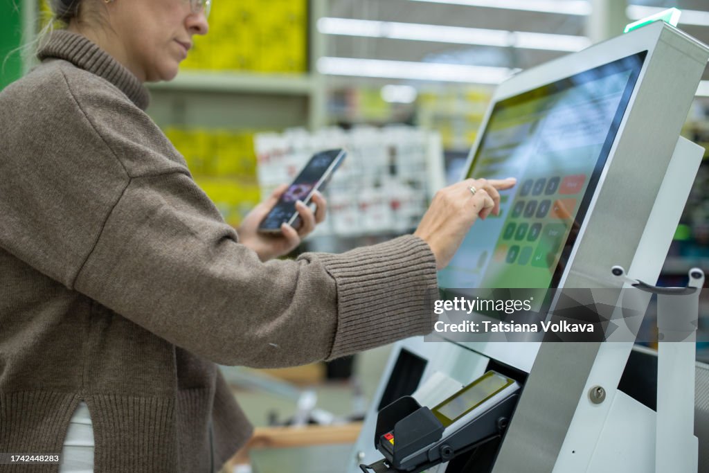 Tech-savvy lady inputs information on device screen while holding smartphone at self-service checkout