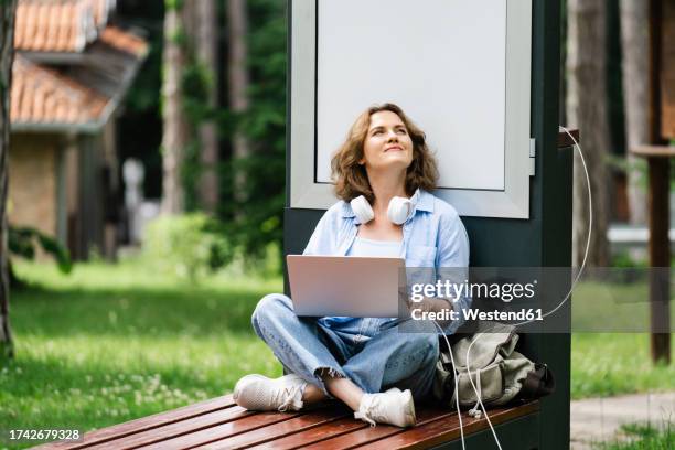 thoughtful woman with laptop sitting near solar charging point at park - cross legged stock pictures, royalty-free photos & images