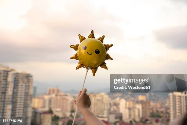 hand of woman holding sun shaped smiley balloon against skyand city skyline - smiley face stock pictures, royalty-free photos & images