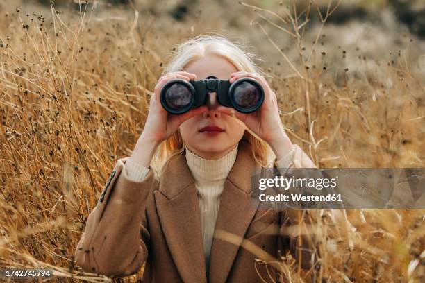 blond woman looking through binoculars in field - paire de jumelles photos et images de collection