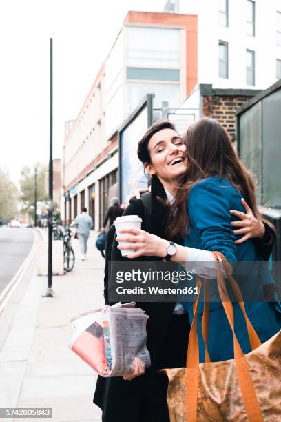 happy businesswoman embracing friend holding coffee cup at sidewalk - long coat stock pictures, royalty-free photos & images