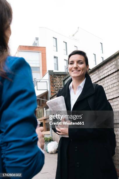 smiling businesswoman holding newspaper and talking with office colleague - long coat stock pictures, royalty-free photos & images