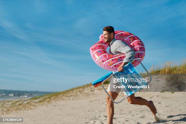happy man running with inflatable swim ring and folding chair at beach on sunny day - folding chair stock pictures, royalty-free photos & images