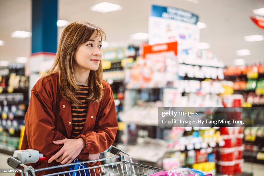 Young pretty Asian woman waiting in line for checkout while shopping for groceries in supermarket