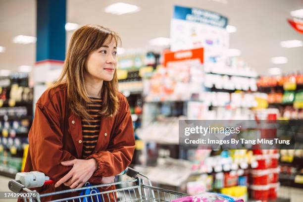 young pretty asian woman waiting in line for checkout while shopping for groceries in supermarket - supermarket queue stock pictures, royalty-free photos & images