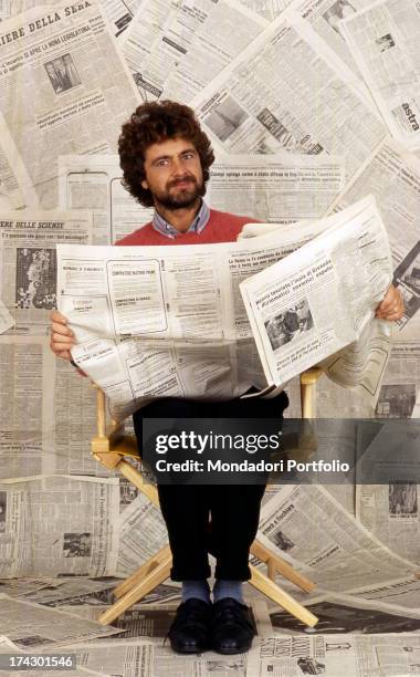 The Italian comedian Beppe Grillo, seated on a folding chair, is holding a newspaper in his hands. Behind him a photographer's limbo covered with...