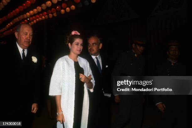 Grace Kelly and the prince Ranieri of Monaco, walk arm in arm, escorted by a person in a black suit and two policemen; the street is brighten by...