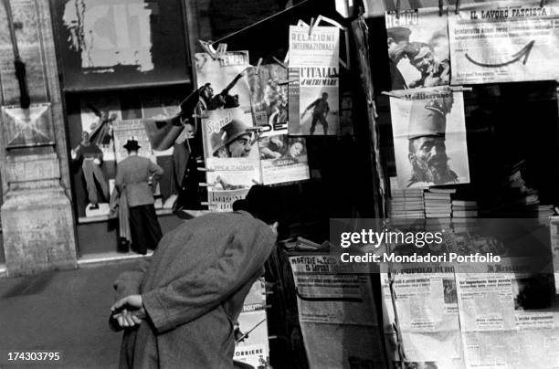 Passerbyer stoops and curiously looks at the headline of a daily newspaper, on display in a kiosk. Italy, 1940..