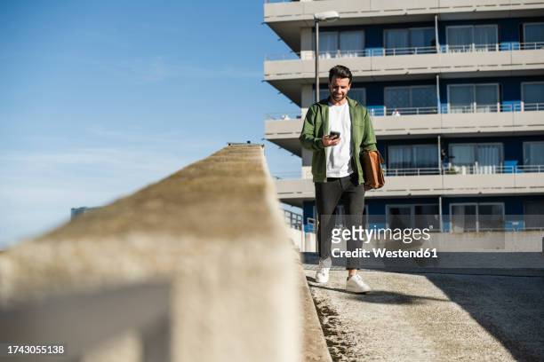 man using smart phone and walking with bag in front of building - facing front stock pictures, royalty-free photos & images